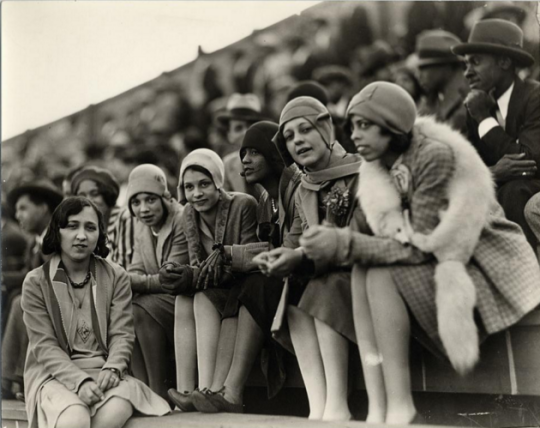 Six black women (Howard University students) in audience, sitting in stadium watching game, men shown sitting behind them. All but one of the women wear cloche hats. The woman on the extreme right was identified as Elise Dowling.