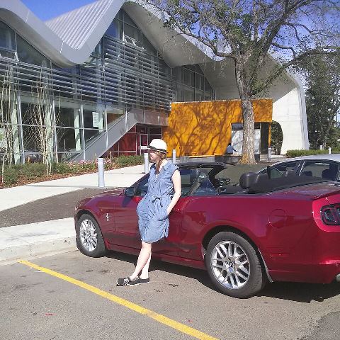 A picture of Jasper Place Public Library in Edmonton Alberta. A red convertible is in the foreground of the image and the author is leaning against the car, wearing a denim dress and a sunhat.